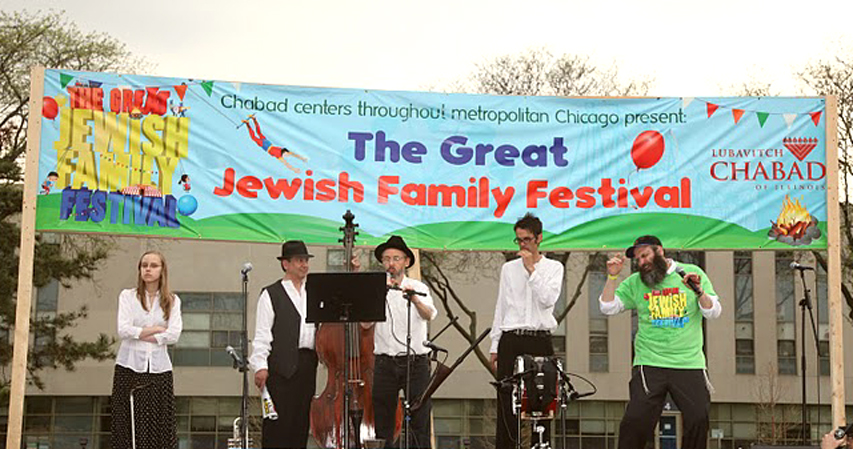 Four musicians of the Ensemble M’chaiya (tm) starting a sound check at the Skokie, IL Chabad festival standing in front of a large banner and next to the Master of Ceremony. © 2011 Modal Music, Inc. (tm) All rights reserved.